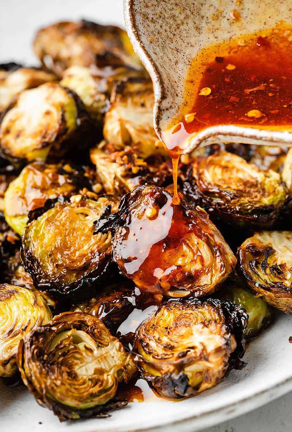 Pouring sauce over a dish of crispy, golden-brown Brussels sprouts. The sauce is drizzling evenly, adding a glossy sheen to the roasted sprouts.