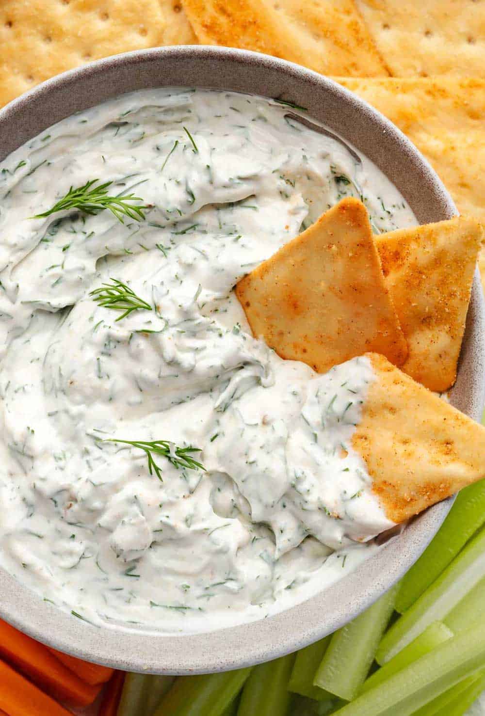 A bowl of creamy dill dip sits at the center, surrounded by an assortment of fresh vegetables, crispy crackers, and golden pita bread chips.