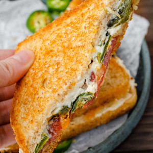 Image of a hand holding a jalapeno popper grilled cheese sandwich, showing the side with melted cheese and visible jalapeno pieces.