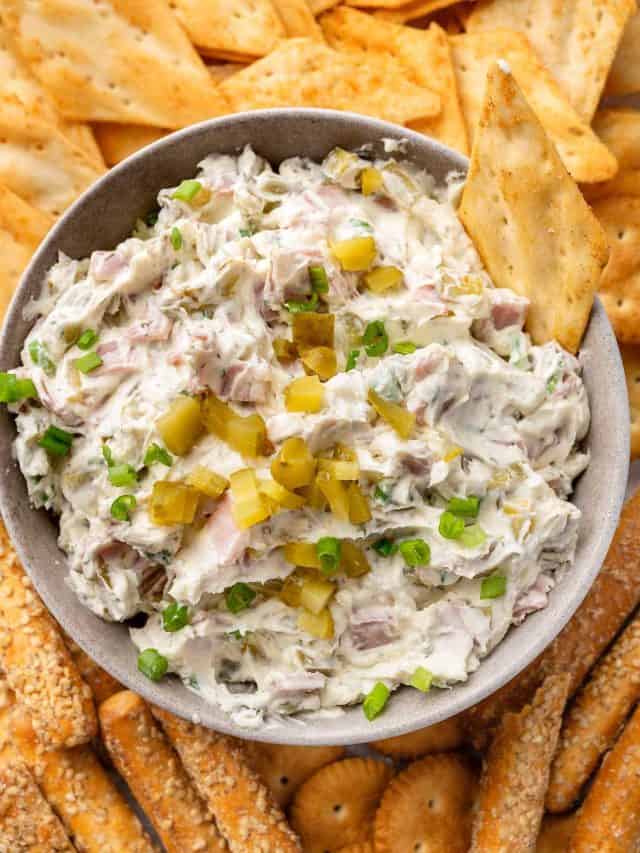 A bowl of creamy dill pickle dip, garnished with chopped pickles, surrounded by pita bread chips and breadsticks for dipping.