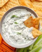 A bowl of creamy dill dip sits at the center, surrounded by an assortment of fresh vegetables, crispy crackers, and golden pita bread chips.