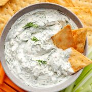 A bowl of creamy dill dip sits at the center, surrounded by an assortment of fresh vegetables, crispy crackers, and golden pita bread chips.