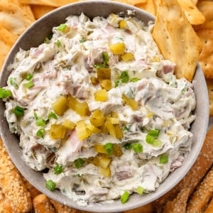 A bowl of creamy dill pickle dip, garnished with chopped pickles, surrounded by pita bread chips and breadsticks for dipping.