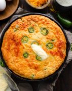 A golden-brown Jalapeno Cheddar Cornbread fresh out of the oven, resting in a cast iron skillet. The cornbread is topped with melting butter.