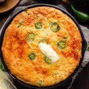 A golden-brown Jalapeno Cheddar Cornbread fresh out of the oven, resting in a cast iron skillet. The cornbread is topped with melting butter.