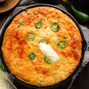 A golden-brown Jalapeno Cheddar Cornbread fresh out of the oven, resting in a cast iron skillet. The cornbread is topped with melting butter.