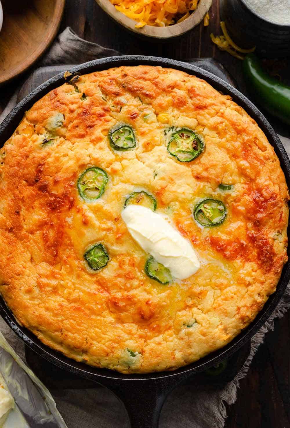 A golden-brown Jalapeno Cheddar Cornbread fresh out of the oven, resting in a cast iron skillet. The cornbread is topped with melting butter