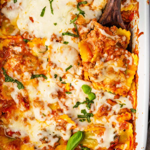 A freshly baked ravioli casserole in a dish, with a serving spoon inside, showcasing layers of ravioli, meaty marinara sauce, and melted cheese on top.