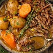 The image shows a rustic bowl filled with tender, shredded chuck roast, whole baby potatoes, and large carrots, garnished with fresh thyme and parsley in a rich, flavorful broth. A spoon rests in the dish, ready to serve.