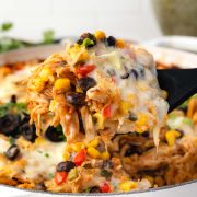 A close-up of a cheesy chicken burrito casserole with black beans, corn, and colorful peppers being scooped out with a spoon.