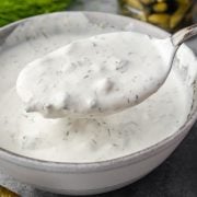 A spoonful of creamy dill pickle ranch dressing is being lifted from a white bowl, with fresh dill and a jar of pickles in the background.