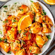 A bowl of white rice topped with crispy orange chicken coated in a glossy, tangy sauce, garnished with black sesame seeds, fresh green onions, and a juicy orange slice.
