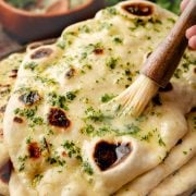 Freshly cooked naan bread brushed with melted garlic butter and sprinkled with chopped herbs, with a bowl of garlic butter in the background.