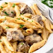 A white bowl filled with creamy Philly Cheesesteak Pasta, featuring penne, tender steak pieces, melted cheese, and fresh parsley, with a fork on the side.