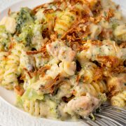 A plate of creamy chicken broccoli pasta casserole topped with crispy fried onions, with a fork digging into the cheesy, golden-brown dish.