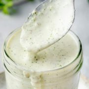A close-up of a spoon lifting creamy jalapeno ranch dressing from a glass jar, with sauce dripping back into the jar.