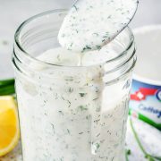 A glass jar filled with creamy homemade ranch dressing with fresh herbs, dripping from a spoon, with a lemon slice and cottage cheese container in the background.