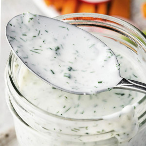 A jar of creamy homemade ranch dressing with fresh herbs, with a spoon resting on top.