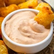 A crispy golden tater tot dipped into a creamy, flavorful sauce, served with a plate of more crunchy tots.