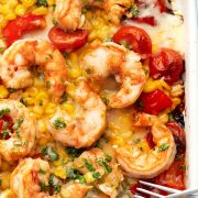 Close-up of baked shrimp with corn, tomatoes, and melted cheese on a white baking sheet.
