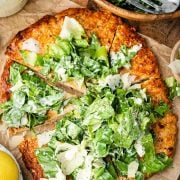 A sliced chicken crust caesar salad pizza topped with fresh with Caesar salad and cheese sits on parchment paper, with salad utensils and a bowl of extra salad nearby.