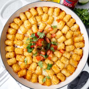 Fiesta Chicken Tater Tot Casserole baked in a white skillet, topped with cheese, salsa, jalapeรฑos, and cilantro.