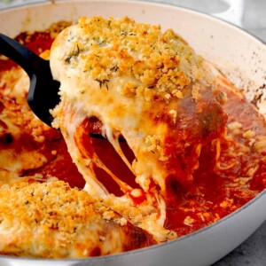 Cheesy chicken parmesan casserole being lifted from a white skillet, showing golden breadcrumb topping and gooey melted cheese with rich marinara sauce.