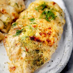 Close-up of juicy melt in your mouth chicken breast baked with creamy Parmesan topping and fresh herbs on a plate.