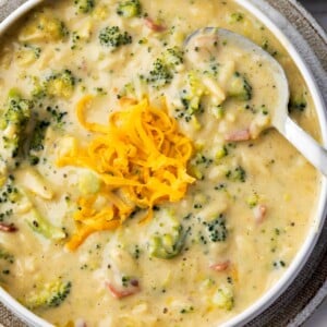Bowl of creamy broccoli cheddar orzo soup topped with shredded cheddar cheese and served with a spoon.