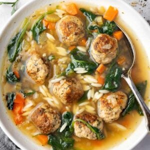A bowl of Italian Wedding Soup filled with tender meatballs, carrots, celery, spinach, and orzo pasta in a flavorful chicken broth, with a spoon resting inside the bowl.