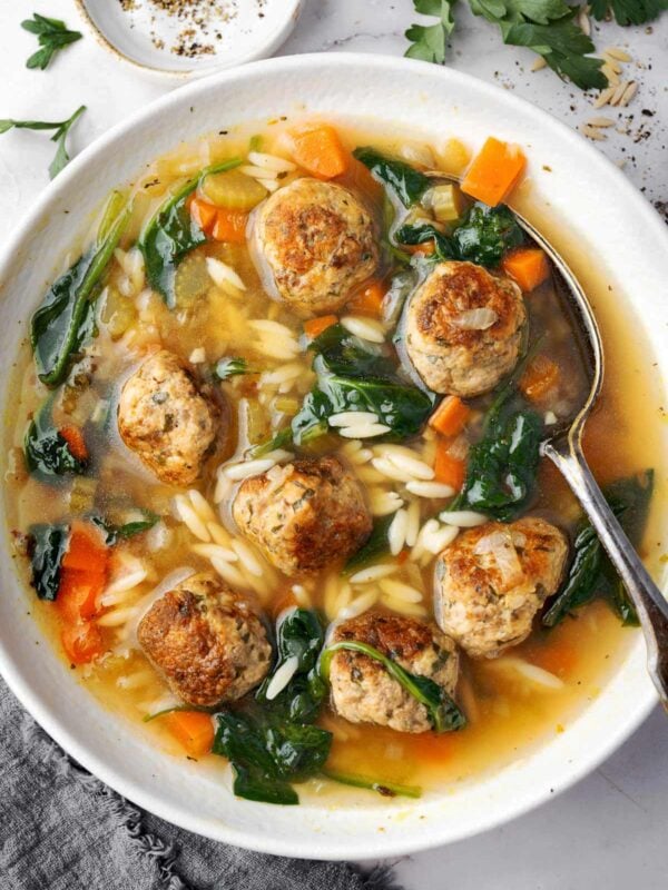 Italian Wedding Soup with Orzo served in a white bowl, filled with homemade meatballs, tender carrots, celery, spinach, and orzo pasta in a flavorful chicken broth, with a spoon and fresh herbs on the side.
