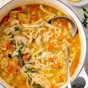 Homemade lemon chicken orzo soup in a white pot with shredded chicken, carrots, celery, and orzo pasta in a golden broth, garnished with fresh parsley and lemon slices on the side.