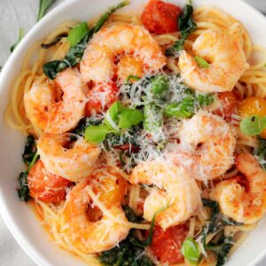 Bowl of pasta with shrimp, cherry tomatoes, and fresh spinach topped with grated Parmesan and basil โ easy shrimp pasta recipe with tomatoes and spinach.