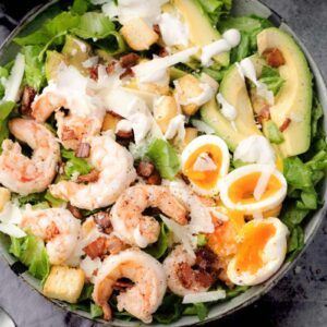 Shrimp Caesar Salad with avocado, soft-boiled eggs, bacon, croutons, and Parmesan, drizzled with creamy dressing.