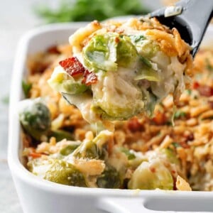 Creamy Brussels sprout casserole with melted cheese and crispy bacon being scooped from a white baking dish.