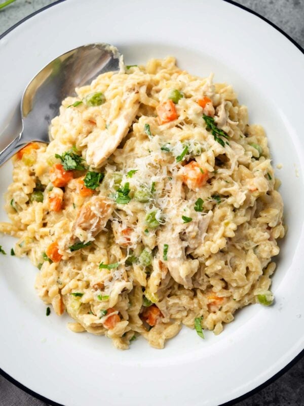 Creamy Chicken Pot Pie Orzo served on a white plate with a spoon, featuring tender chicken, carrots, peas, and orzo pasta topped with grated parmesan and fresh parsley.