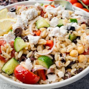 Greek Orzo Salad served in a white bowl with cucumbers, cherry tomatoes, Kalamata olives, red onion, chickpeas, feta cheese, and lemon slices.
