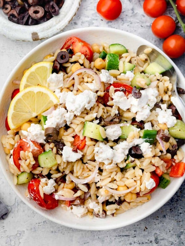 Greek Orzo Salad in a white bowl with cucumbers, cherry tomatoes, Kalamata olives, red onion, chickpeas, feta cheese, and lemon slices โ a fresh Mediterranean pasta salad perfect for summer.