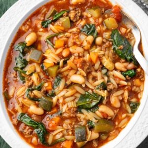 Bowl of hearty minestrone soup with Italian sausage, orzo pasta, white beans, carrots, zucchini, and spinach in a rich tomato broth, served with a spoon.