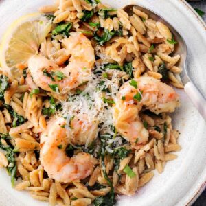Close-up of shrimp scampi with orzo pasta, fresh spinach, grated Parmesan, and lemon slice served on a ceramic plate with a spoon.