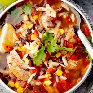 Mexican chicken orzo soup in a bowl with shredded chicken, black beans, corn, tomatoes, cilantro, and lime, served with a spoon on a stone plate.