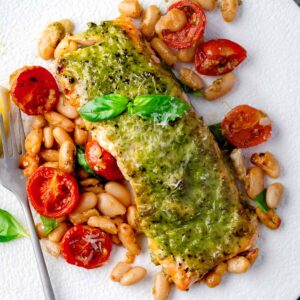 Pesto roasted salmon fillet served with roasted cherry tomatoes, creamy white beans, and fresh basil on a white plate.