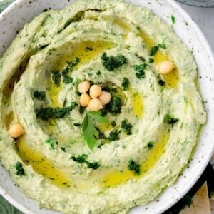 Spinach artichoke hummus topped with olive oil, chickpeas, and fresh herbs, served with pita bread and carrot sticks.