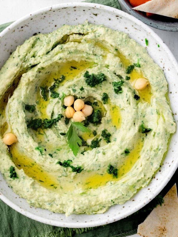 Spinach artichoke hummus topped with olive oil, chickpeas, and fresh herbs, served with pita bread and carrot sticks.
