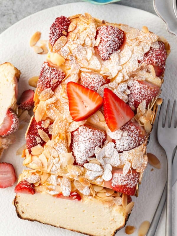 Slice of strawberry cottage cheese breakfast bake topped with fresh strawberries, sliced almonds, and powdered sugar, showing a creamy baked interior served on a white plate.