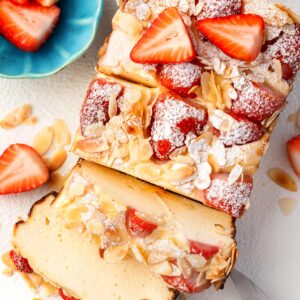Overhead view of strawberry cottage cheese breakfast bake sliced on a plate, topped with fresh strawberries, sliced almonds, and powdered sugar, with extra strawberries on the side.