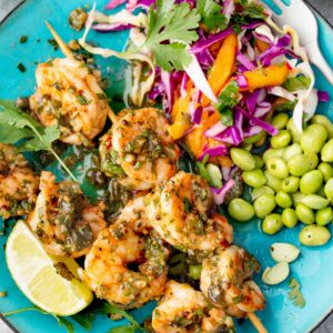 Air fryer jalapeรฑo cowboy butter shrimp skewers served with mango coleslaw, edamame, fresh cilantro, and lime wedges on a blue plate.