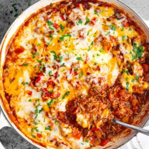 Unstuffed peppers casserole in a white baking dish topped with melted Monterey Jack cheese, ground beef, rice, diced red and yellow bell peppers, rich tomato sauce, and fresh parsley, with a spoon scooping a serving.