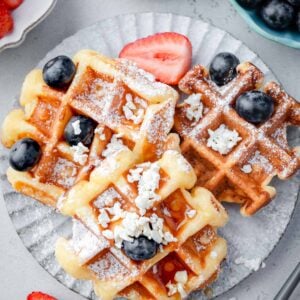 Overhead view of cottage cheese waffles topped with blueberries, powdered sugar, crumbled cottage cheese, and honey, served with fresh strawberries on a plate.
