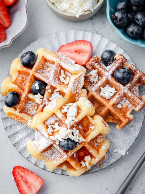Overhead view of cottage cheese waffles topped with blueberries, powdered sugar, crumbled cottage cheese, and honey, served with fresh strawberries on a plate.
